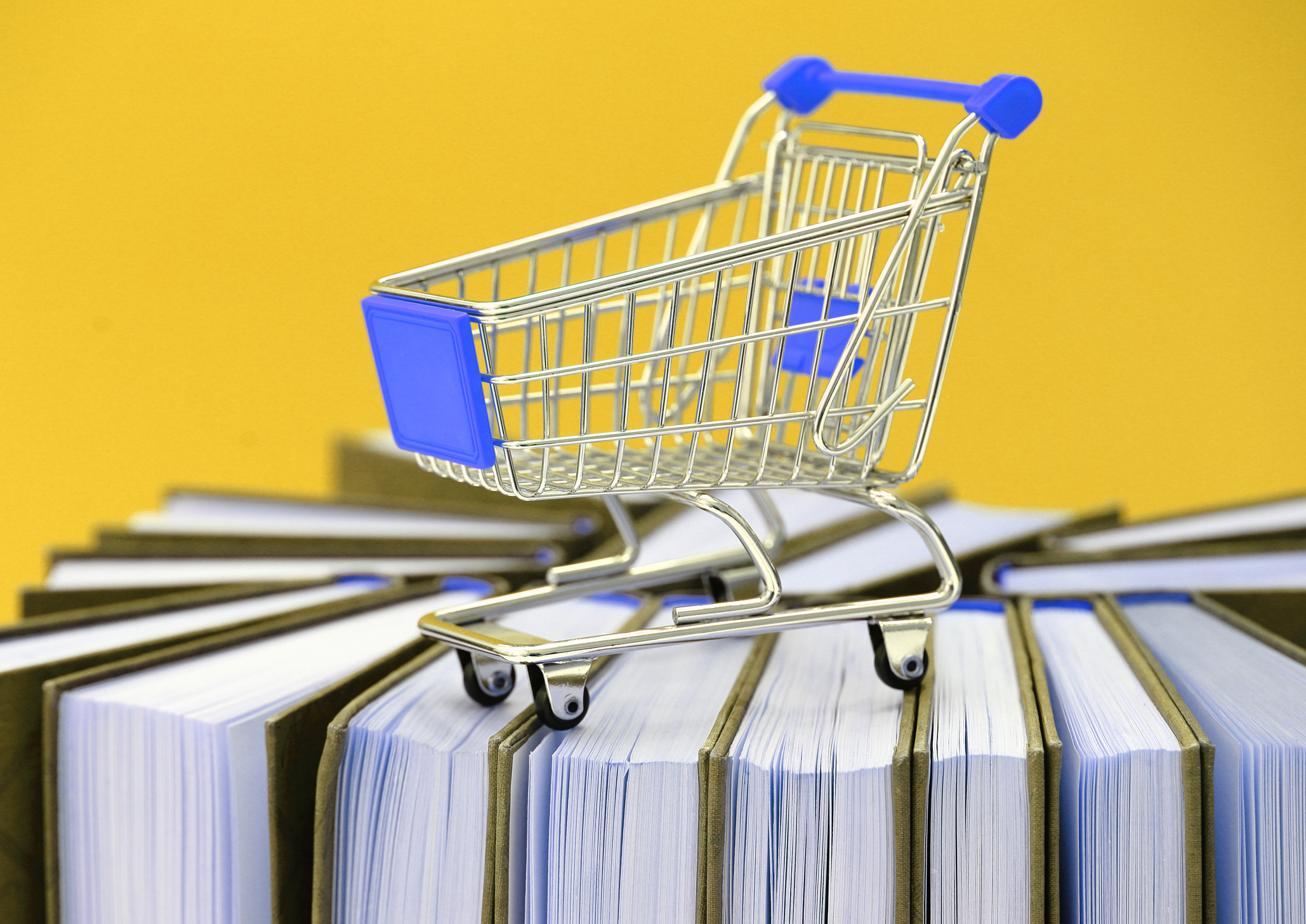 A toy-sized shopping cart is sat on top of a stack of vertical standing books.