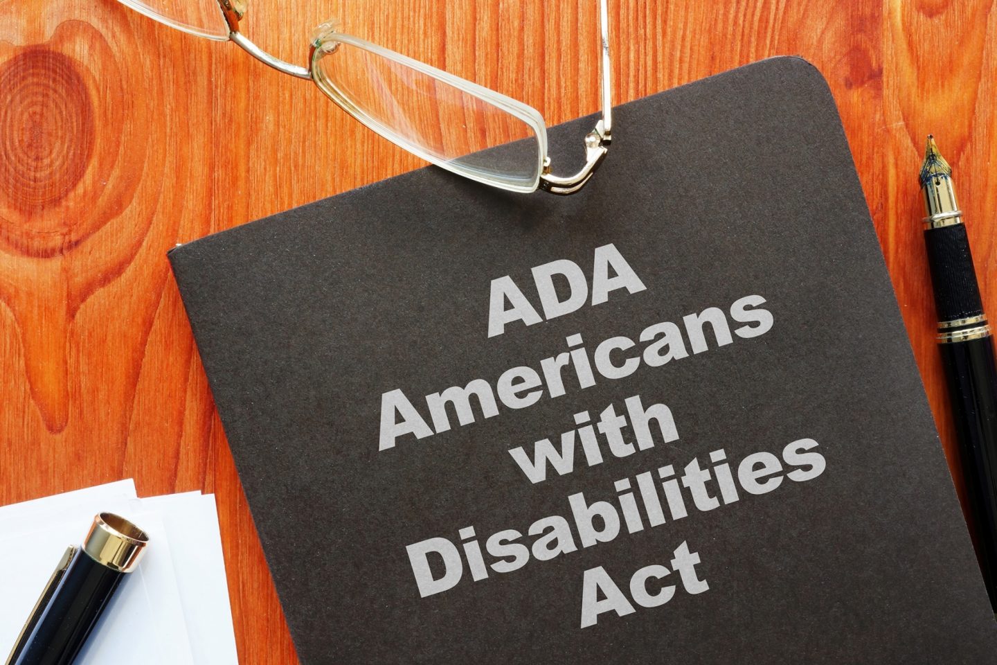 A black book that has "ADA, Americans with Disabilities Act" on the cover. The book sits on a wooden table with a pair of glasses and an open fountain pen surrounding the book.