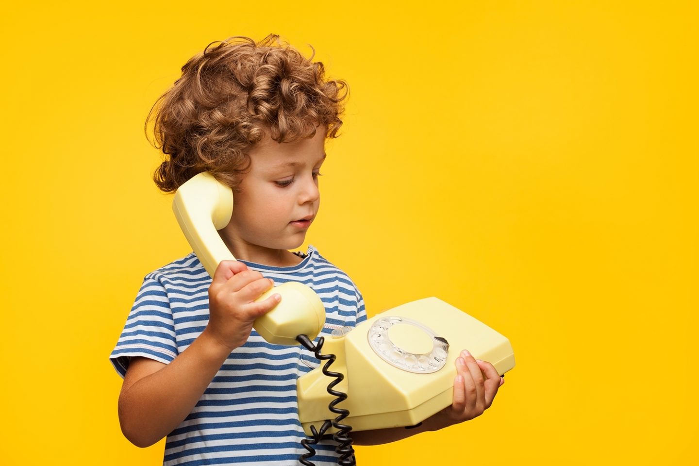 A young curly-haired boy holds a retro rotary dial phone to his ear.