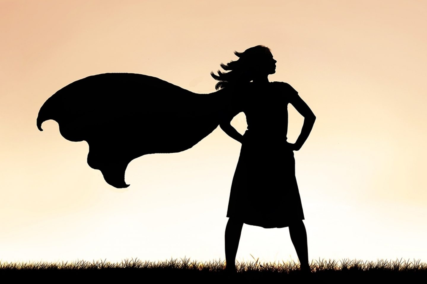 A caped Super Hero Woman stands in grass. She is in all black and is depicted by her silhouette.
