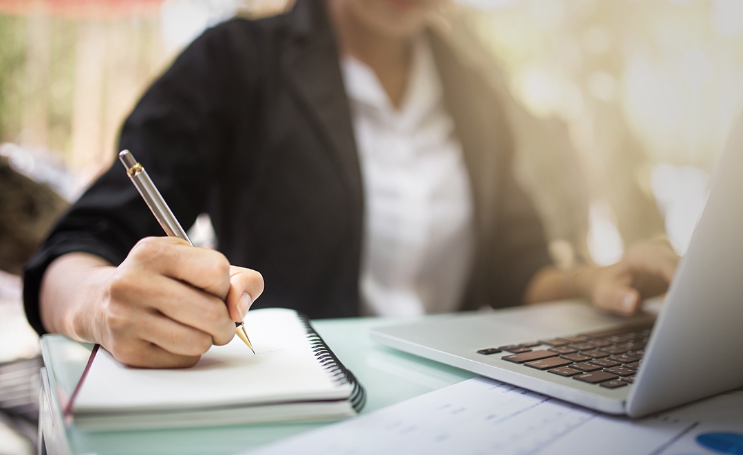 ESOL class A woman uses her laptop with her left hand while taking notes in her notebook with her right hand.