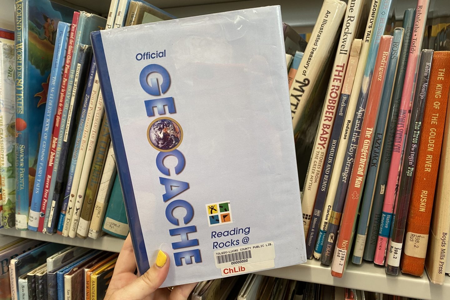 The "Official Geocache" book is held up in front of other books on a shelf in the Children's Library.