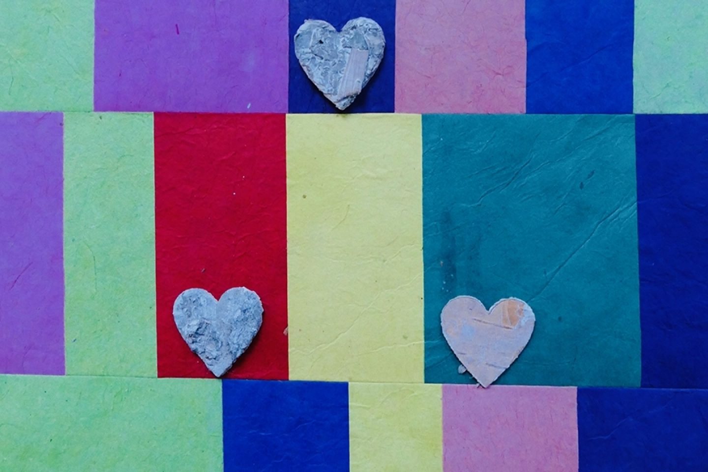 Multi-colored pieces of paper have three wooden hearts placed over the paper in a triangle shape.