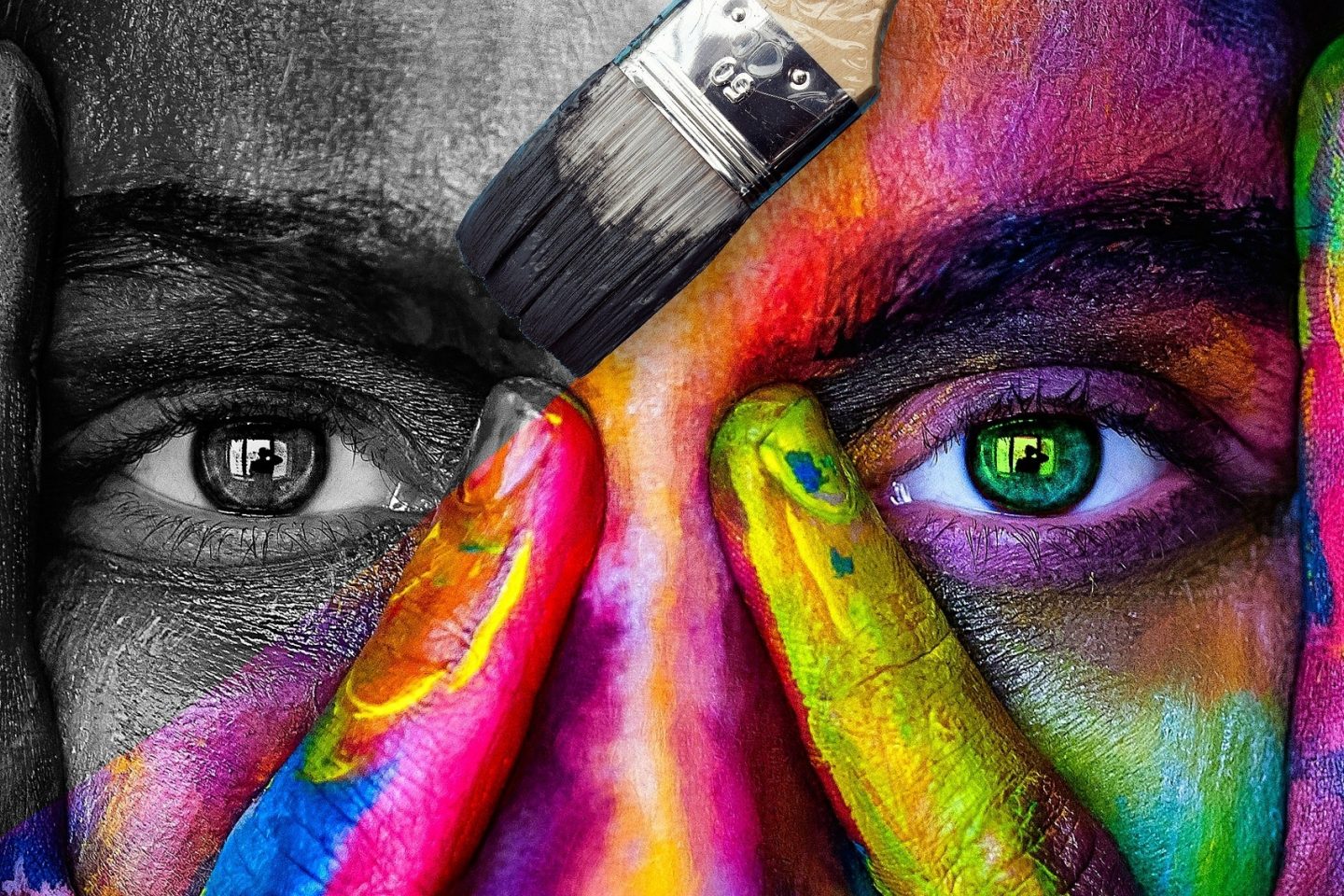 A paintbrush with black paint is coloring a close-up view of a woman's face. Her eyes and part of her nose are visible through her fingers that cover either side of her nose. The brush paints over the left side of the image to be black and white. The right side of the image is full of various bright colors.