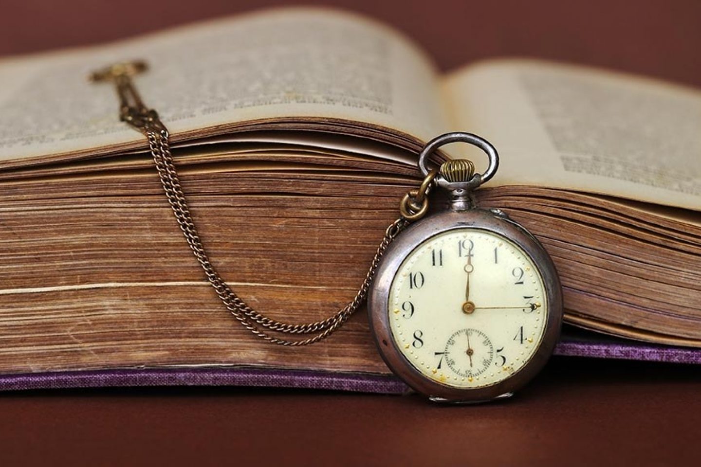 A pocket watch sits in front of an open book. Its face reads "12:15."