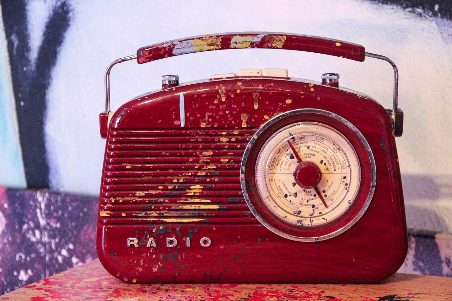 An old red portable radio sits atop a table with yellow paint splatters covering its surface.