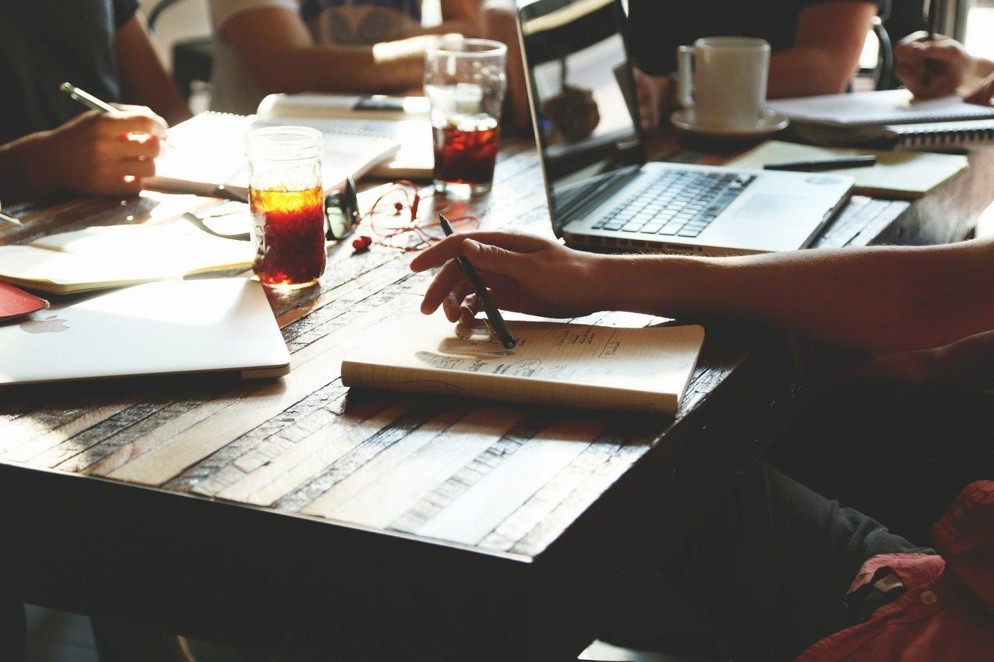 A group of people sit around a table. The view is a close-up of the table, the people's hands, several notebooks, a laptop, and various drinks that surround them.
