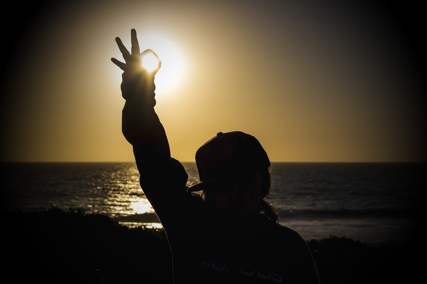 A child has their hand raised and forms the "ok" symbol with their fingers. Their back is to us as they stand in front of a view of the sun. The sun is a bright yellow color and leaves the rest of the image in shadows.