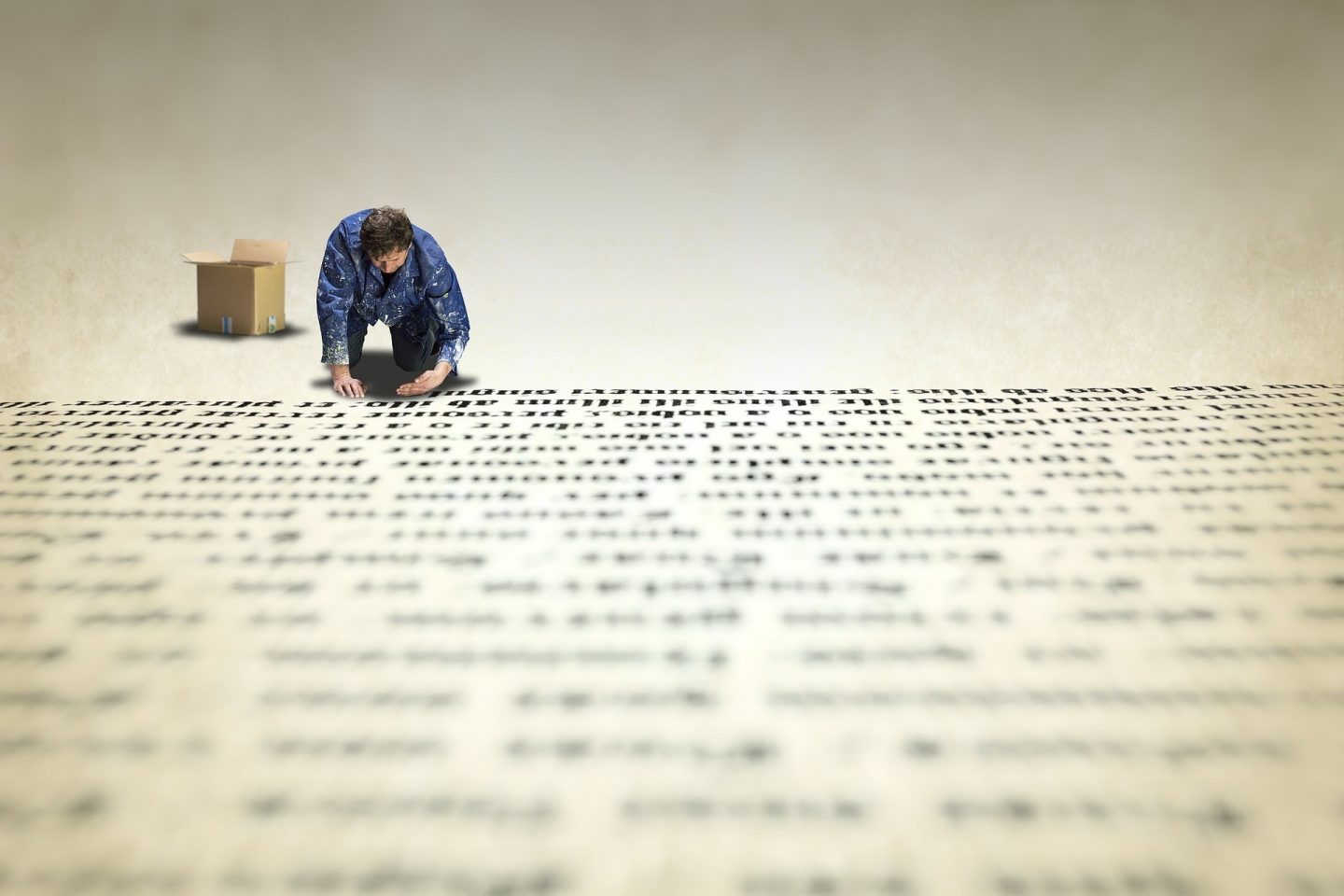 A man is on his knees and uses his hands to place text on the floor. Next to him is a cardboard box where he grabs each new letter to form the words on a page.