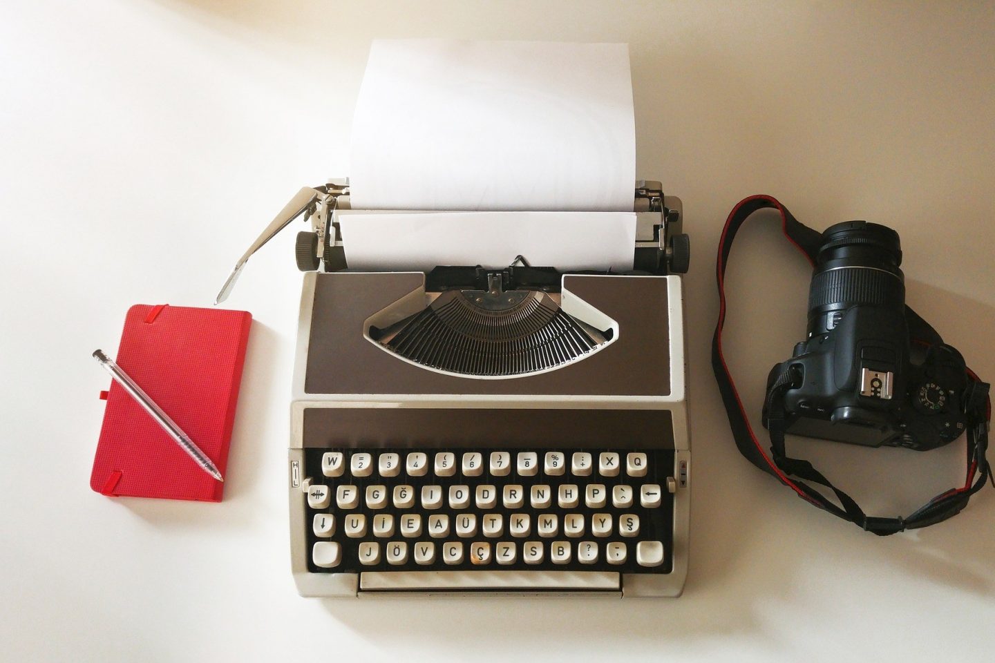 A red notebook with a pen on top of it is left of a typewrite and a digital camera on a table.