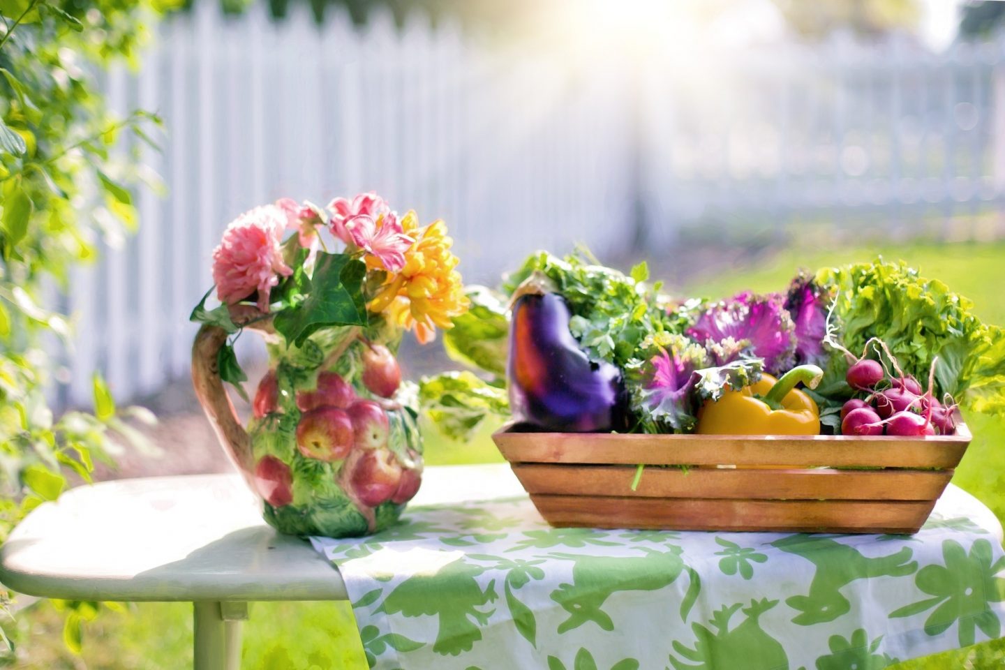Vegetables in a wooden carrier sit on a table next to a vase. The vase is in the shape of a bushel of apples. The vase also has pink and yellow flowers coming out of its opening.