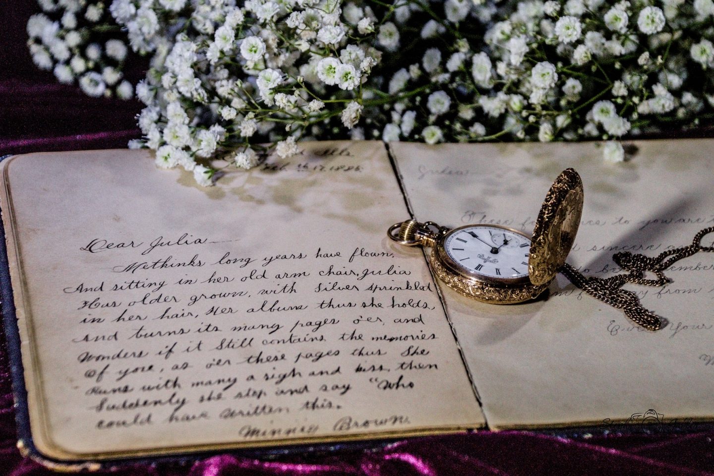 A handwritten letter is left open on a table with a golden pocket watch and vintage flowers laid overtop of the letter.