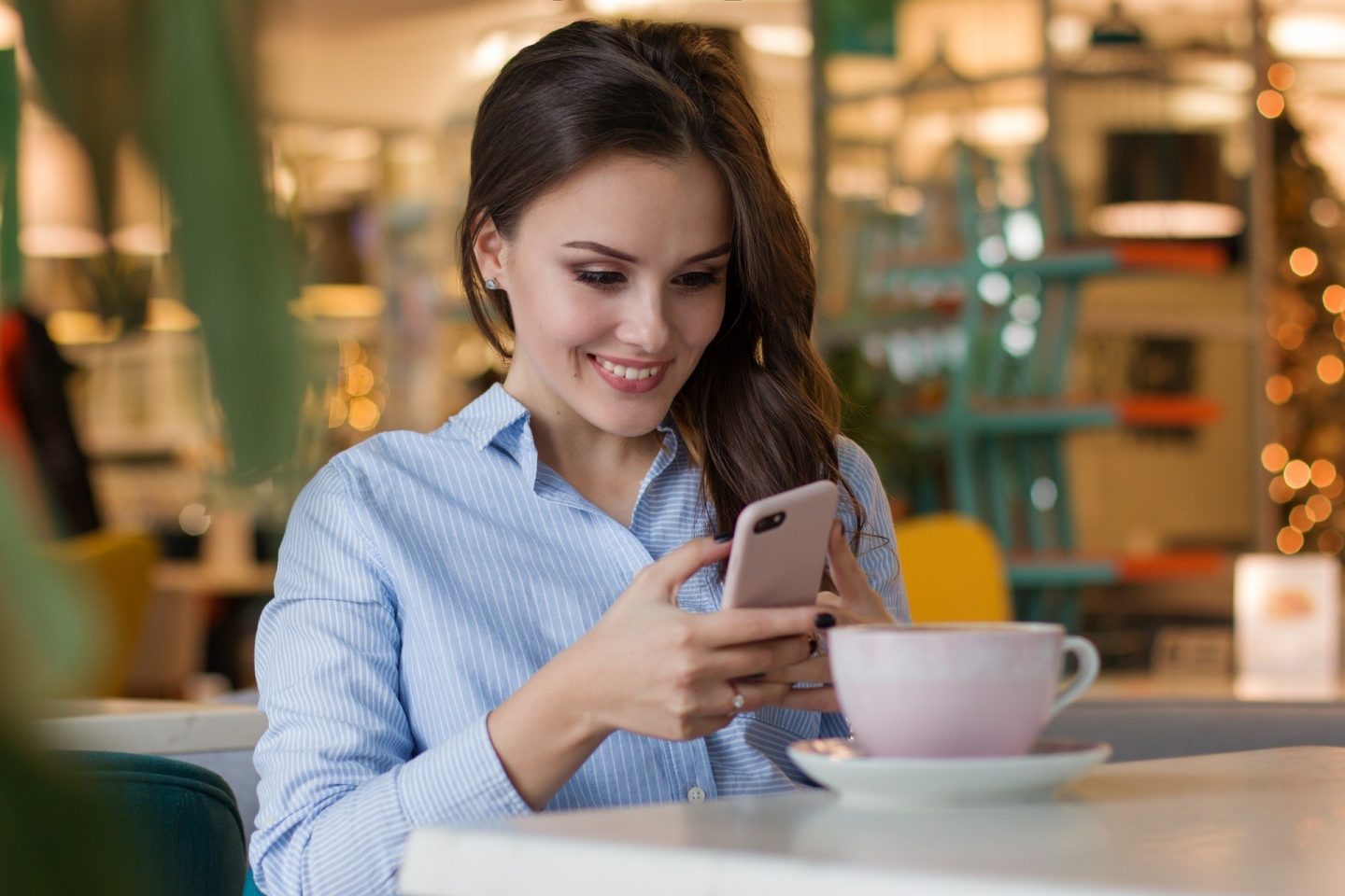 A woman is smiling while using her phone at a coffee shop.