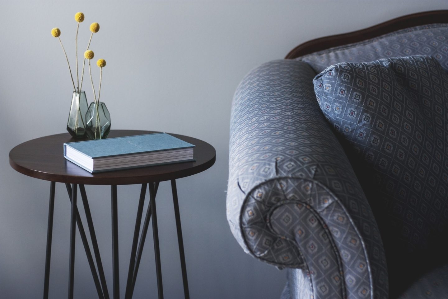 A circular end table holds a blue book and two vases with circular yellow plants. Next to the table is part of a couch's arm and pillow.