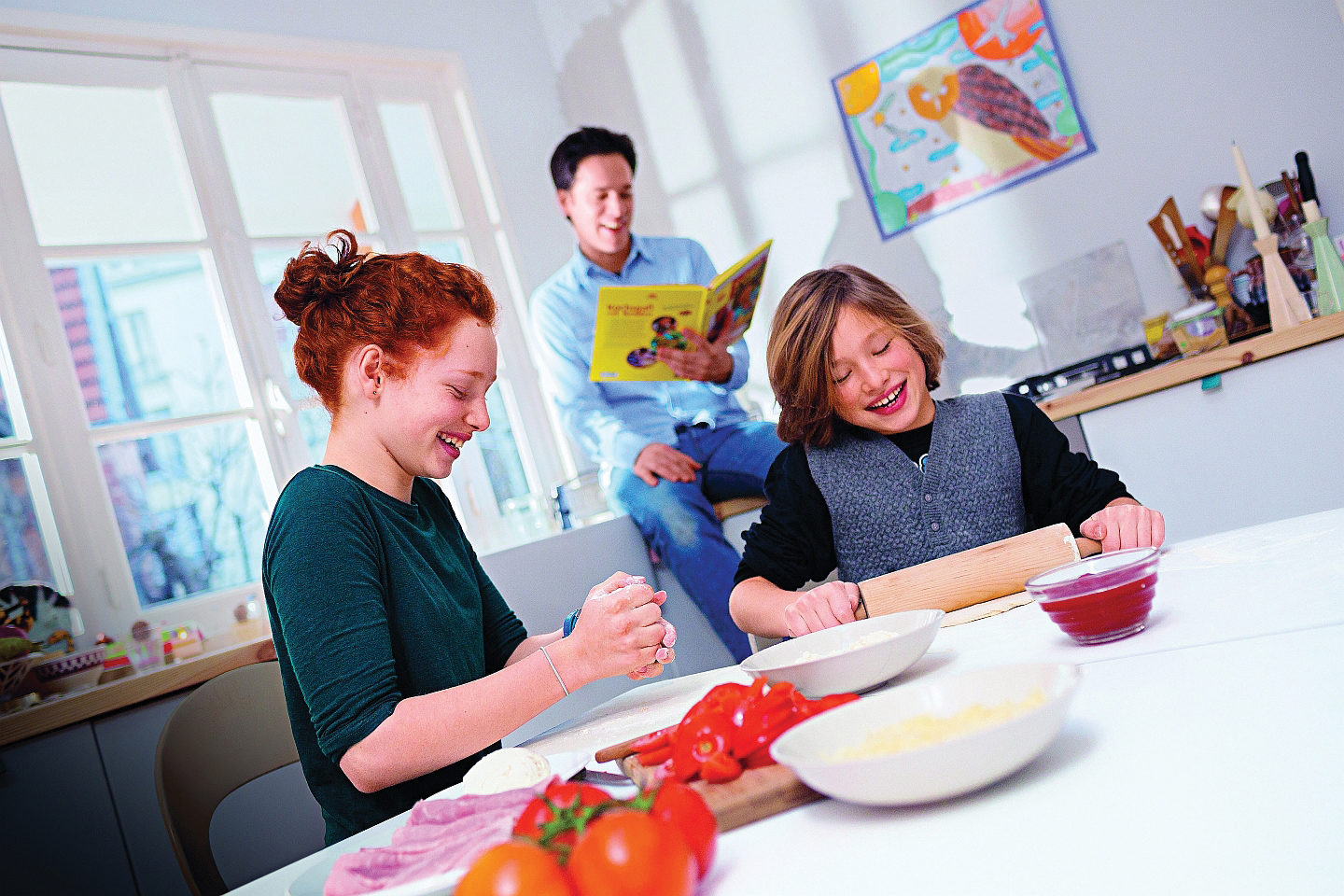 A father sits on a counter top reading a book behind his two children. His two children are sitting at the kitchen table, laughing while making dough for a pizza.
