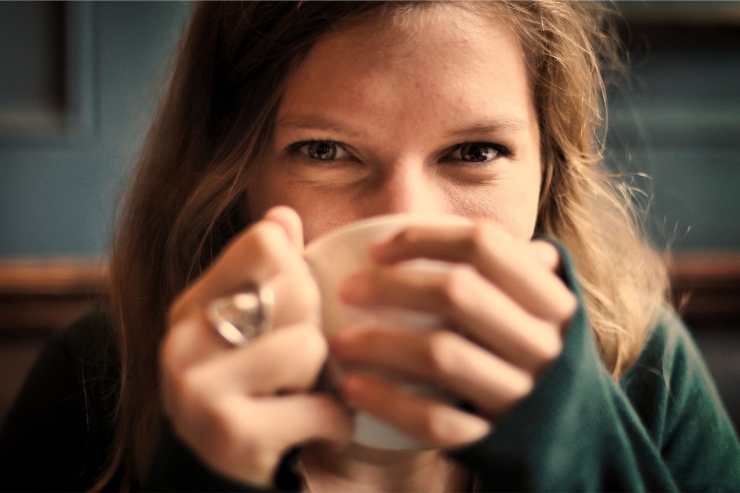 A woman looks forward at the photographer as she holds a mug to her face, obscuring the view of her mouth and nose. She wears a green long sleeve shirt and silver ring on her right hand's middle finger.