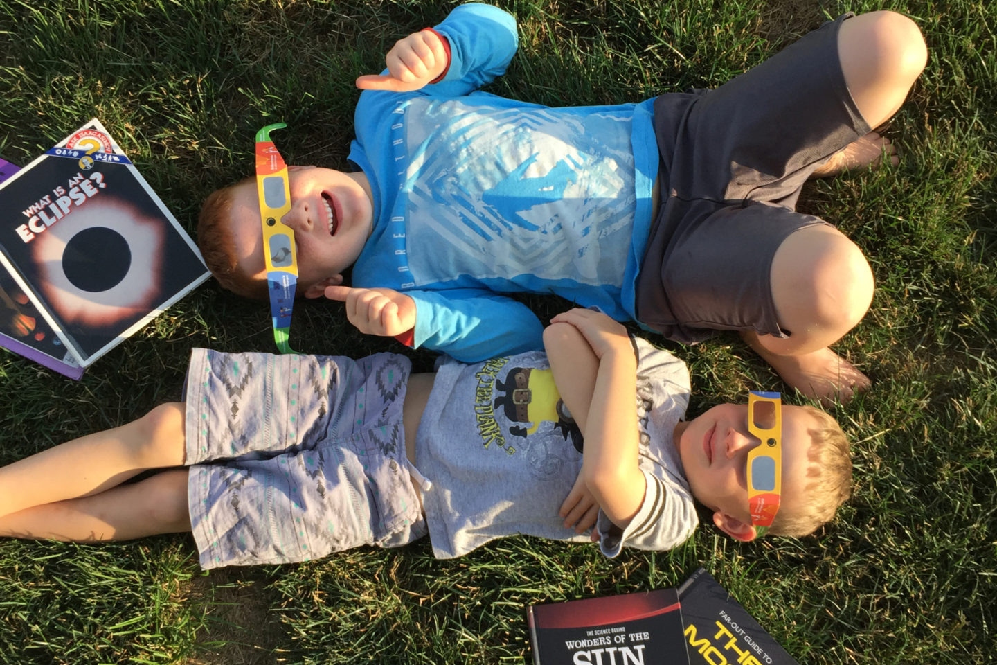 Two young boys lay in the grass on their backs. Both are wearing solar eclipse glasses and have books around them with titles like "What is an Eclipse?" and "Wonders of the Sun."