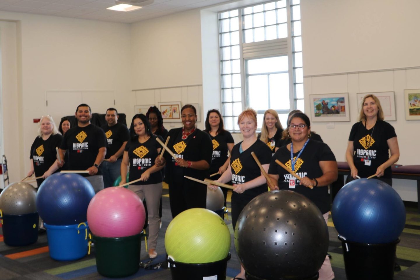 A group photo of the Latino Cultural Committee at the Toledo Lucas County Public Library. Together, the group wears matching T-shirts while they hold drum sticks. In front of each member is an exercise ball inside of a large tub that they use the drum sticks to hit.
