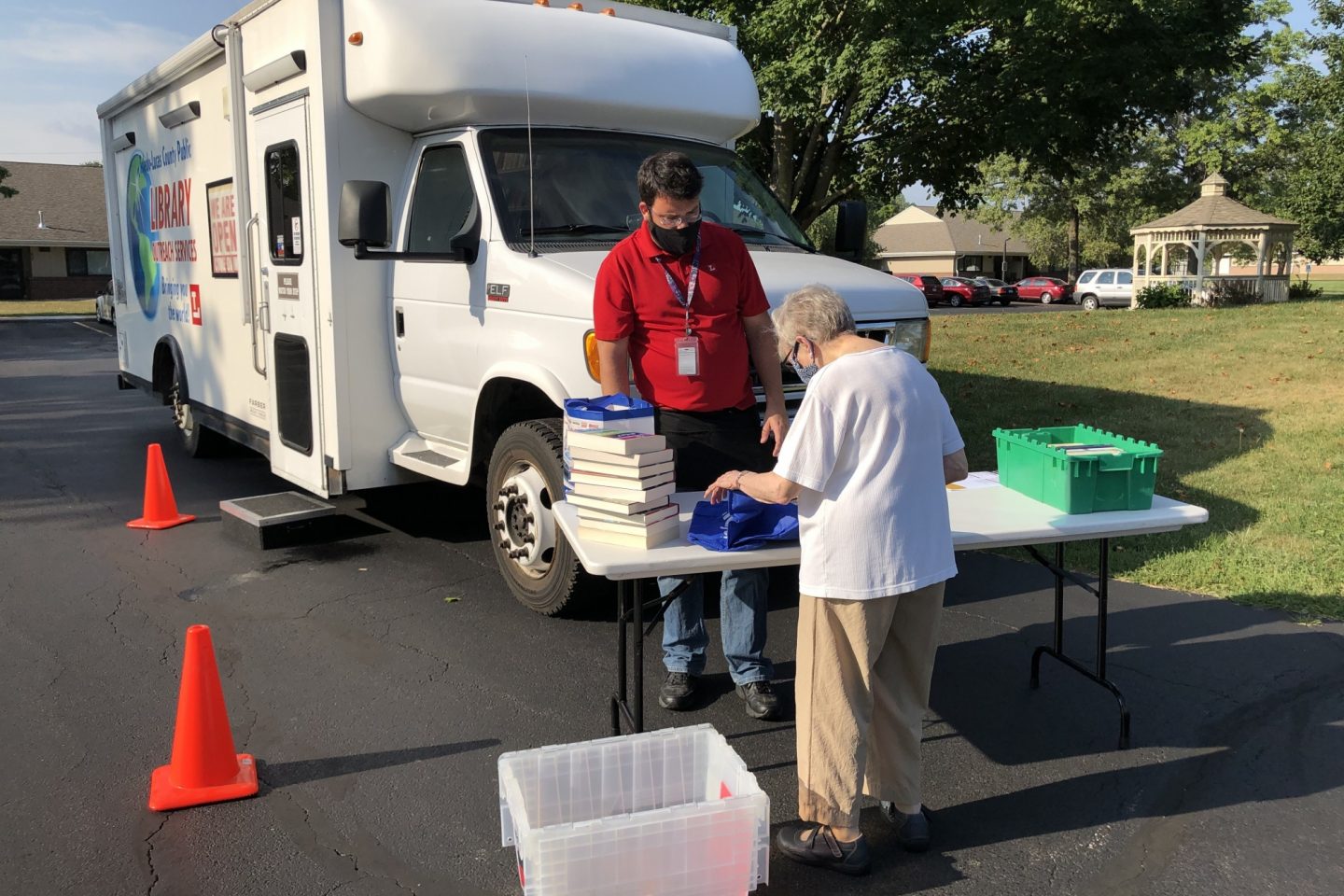 Toledo Lucas County Public Library's Mobile Service unit is out and about in the community. A library worker is stationed at a table aiding an older woman fill her bag with books. Around them is a pile of books and plastic totes that help Mobile Service's workers move materials on and off the bus.