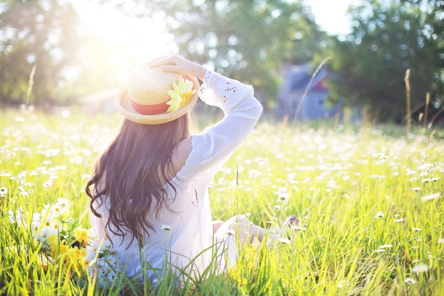 A girl in a white dress with a wicker hat and yellow flower in it sits in a grassy field with the sun illuminating her.