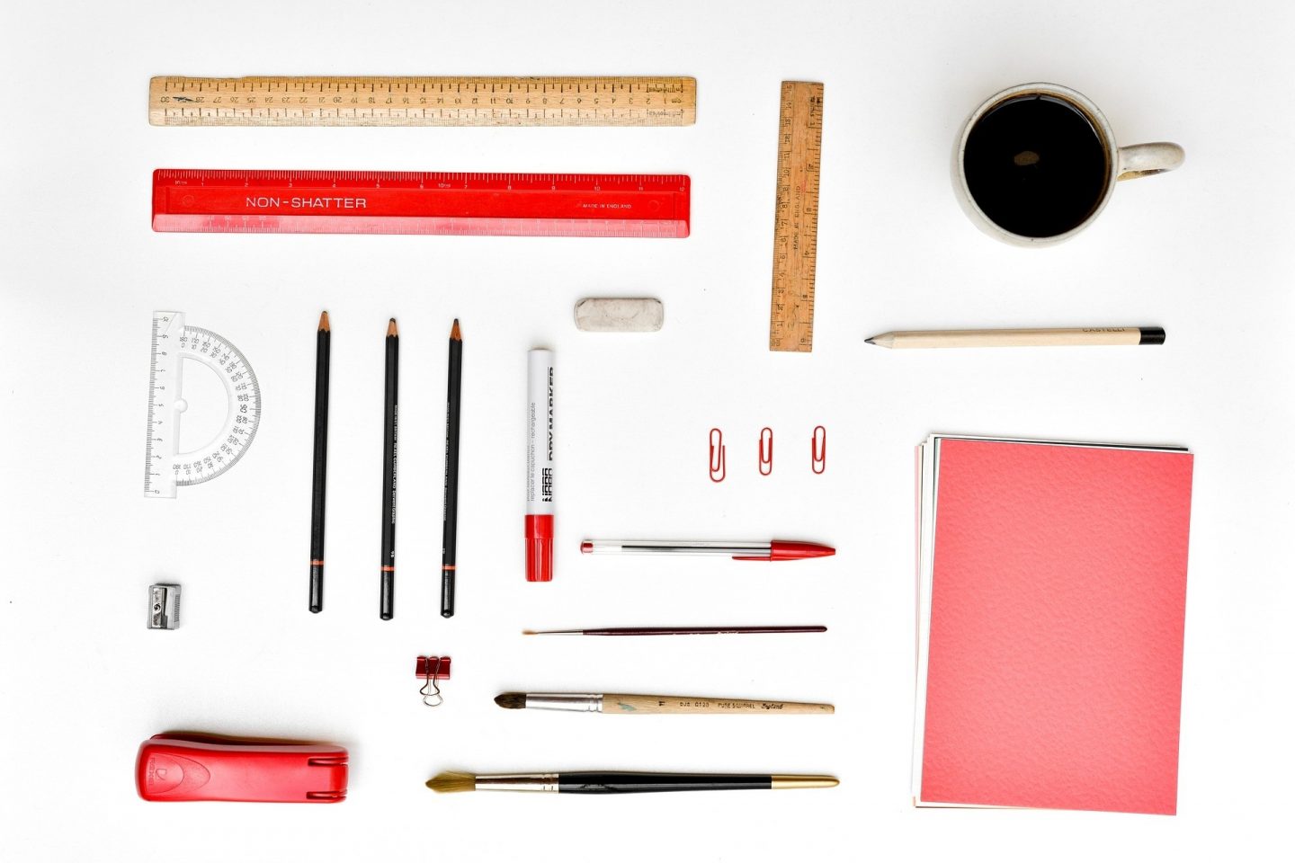 OCD desk with three rulers lines up, several pens, markers, pencils, brushes, and papers. There is also a coffee cup, stapler, and pencil sharpener.
