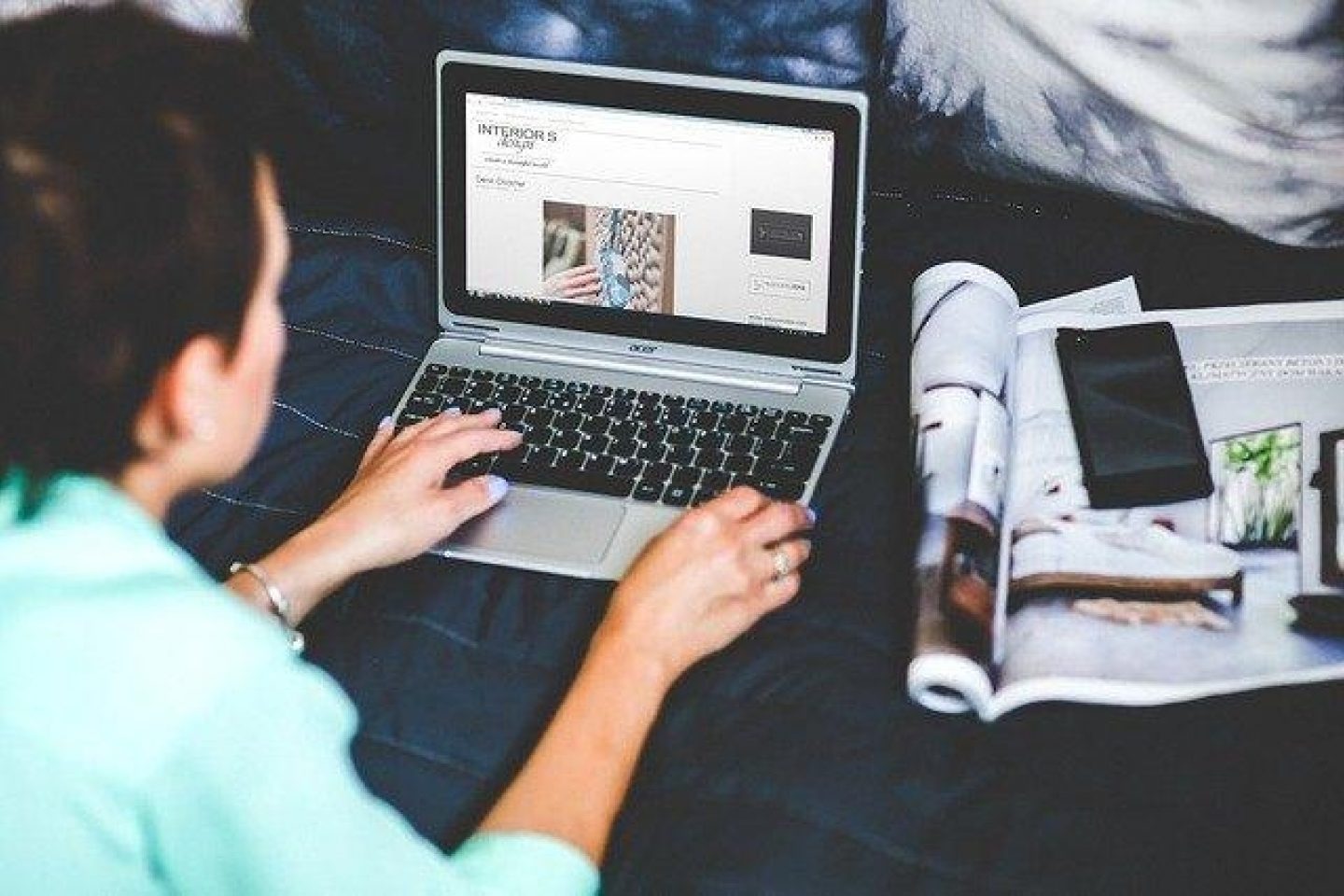 A woman lays on a bed near the pillows with her laptop out and open. She is seen looking at interior designs on the computer, and to her right is a cellphone and magazine also about interior design.