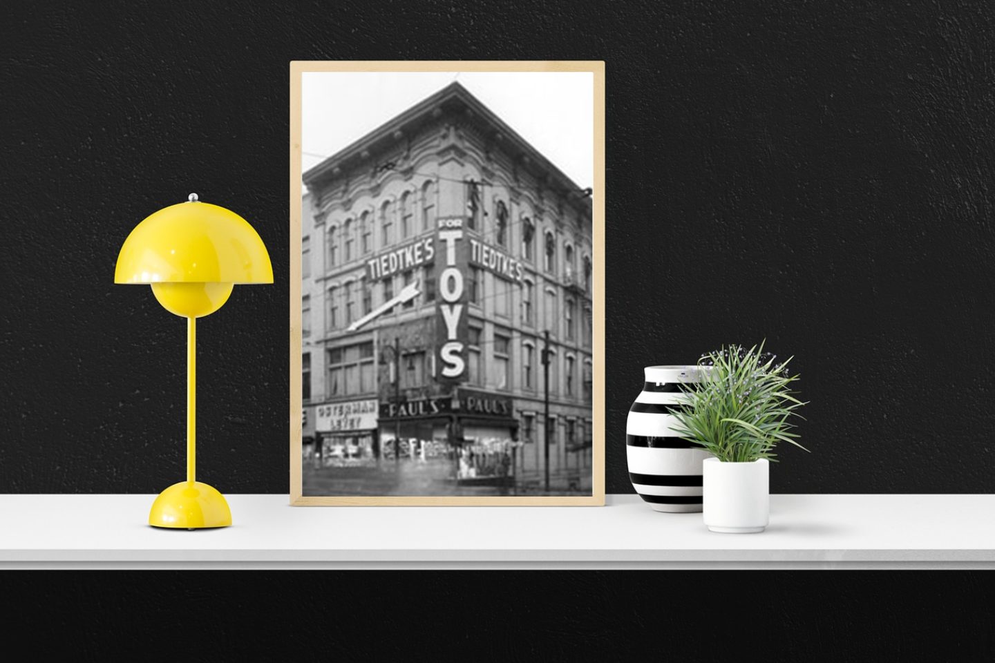 A black-walled room has a white table with a yellow desk lamp and two planters. In the center of the table is a framed black-and-white image. The image in the frame shows a view of the Tiedtke's Store for Toys from the Local History and Genealogy Department.