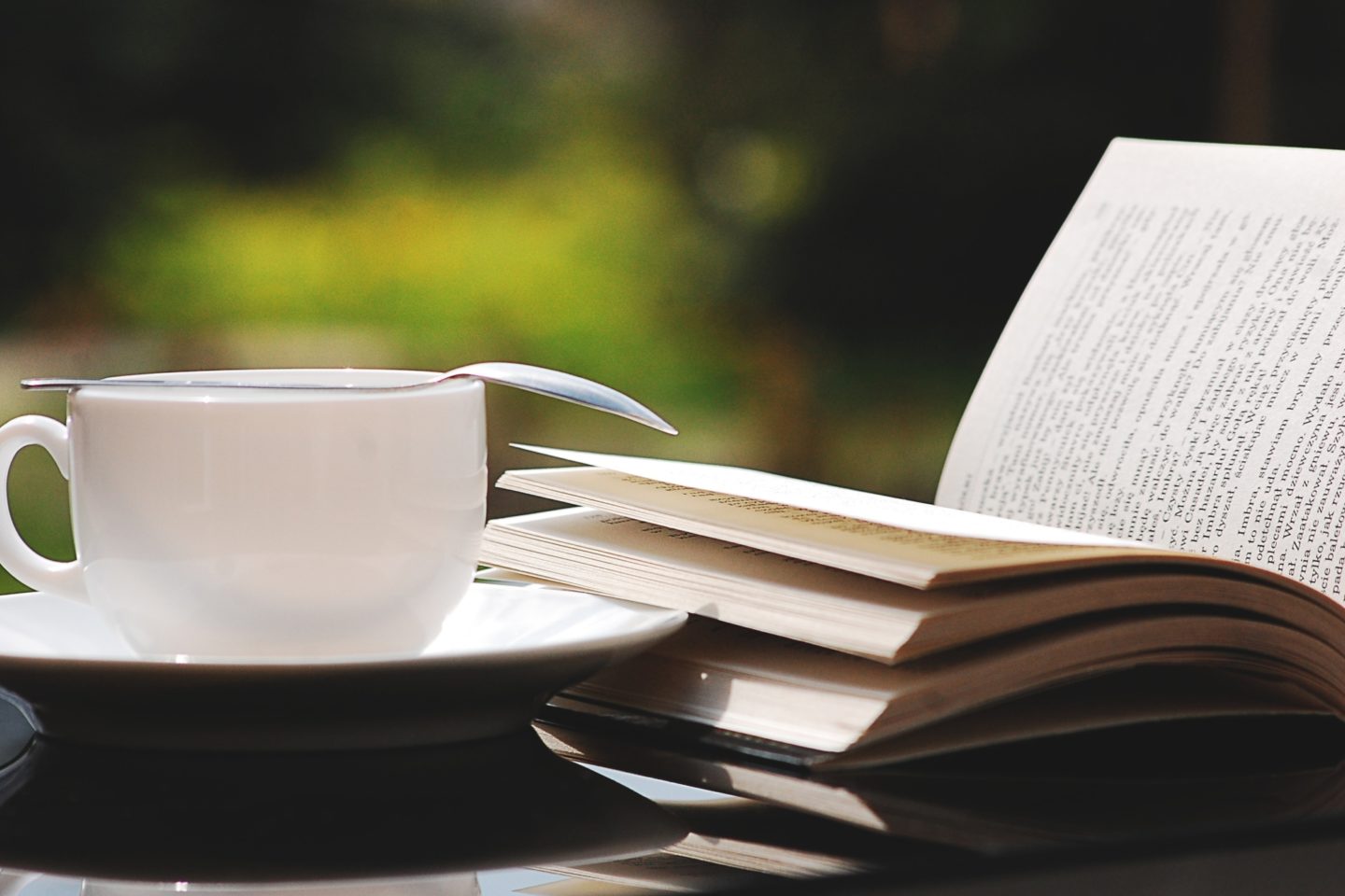 A book is open on a table next to a white mug and a dish. On top of the mug is a silver spoon.