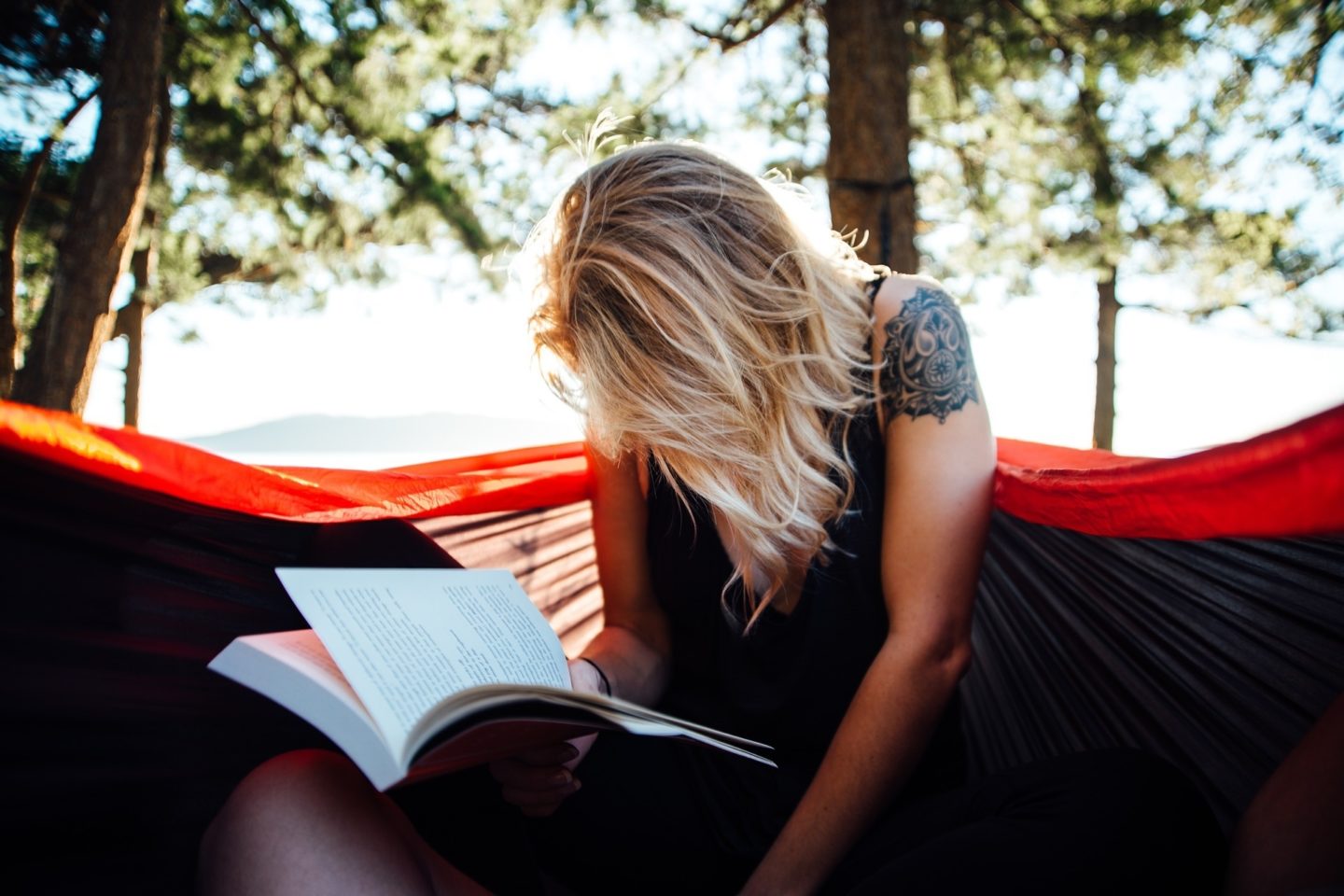 A woman sits in the center of a hammock tied high in a tree. She looks down at the book she is reading, while her hair hides her face. Her left arm has a black tattoo near the top of her shoulder, and behind her is a view of other tall trees and mountains in the distance.