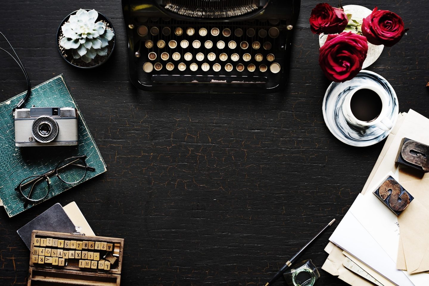 A vintage black typewriter sits on a desk. On either side of it are plants. The left side has a small succulent while the right side has three roses in a glass. The left side of the scene has a camera and black-rimmed glasses on a green journal. Below that are wooden squares with letters, numbers, and symbols on them. The right side of the scene shows a mug with coffee in it on a dish. There are also two large woodblock letters of "S" and "U" on top of manilla envelopes.