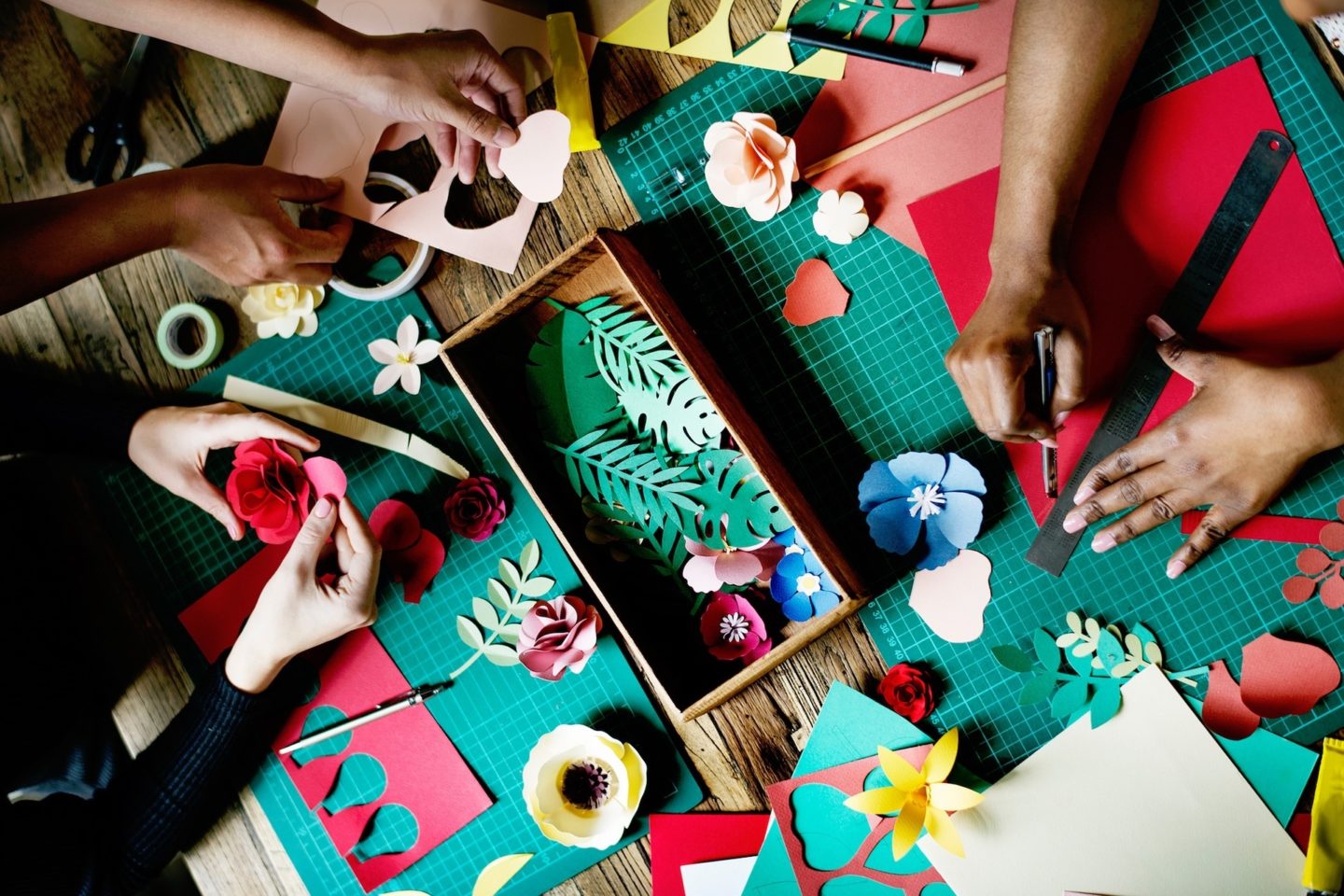 Arts and crafts gift ideas made with paper. Three sets of hands are viewed from above as they work on making different flowers and plants out of paper.