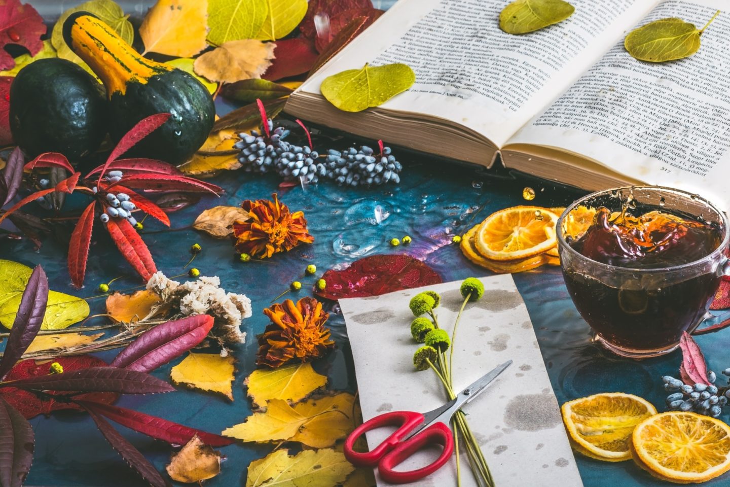 Autumn-themed still life featuring an open book with leaves on the pages, surrounded by colorful fall leaves, two gourds, clusters of berries, dried orange slices, flowers, and a pair of red scissors on a table. A cup with liquid splashing out is also present, creating a vibrant and dynamic scene.