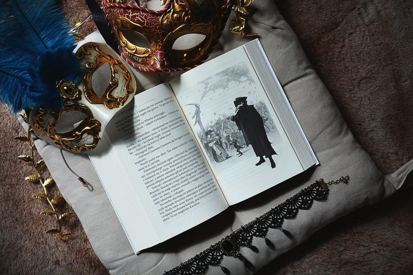 A book sits open on top of a pillow. Surrounding it are two decorative Carnival masks and a black necklace choker. The book is open to a scene depicting Carnival.