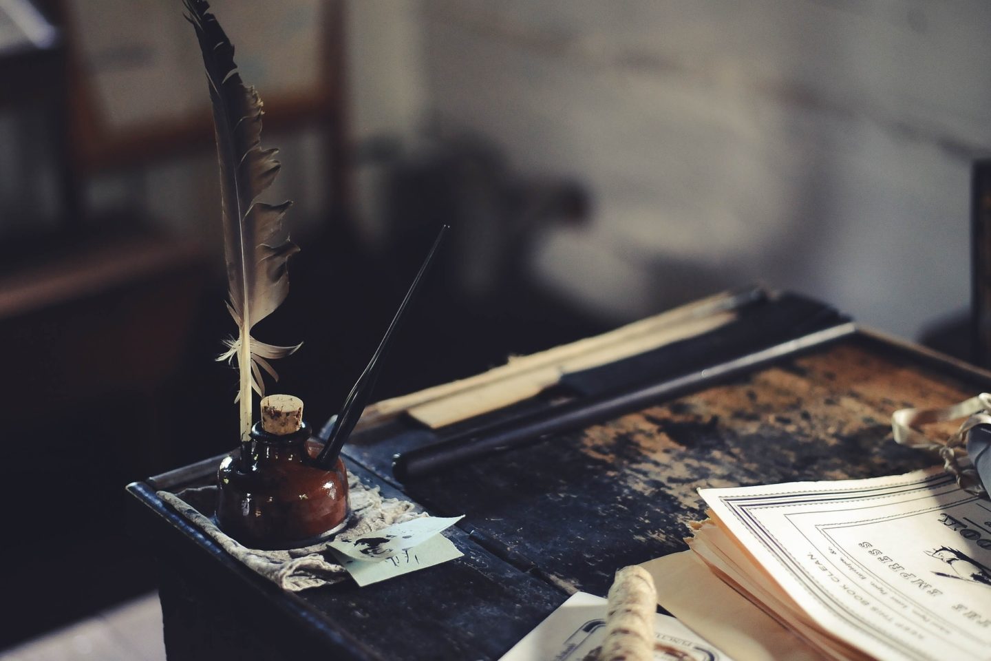 A desk holds a writing quill, ink bottle, and fountain pen on it. Also on the desk is a scrap piece of paper that has some of the ink blotted onto it, along with some papers that appear to have been written with the ink and feather or fountain pen.