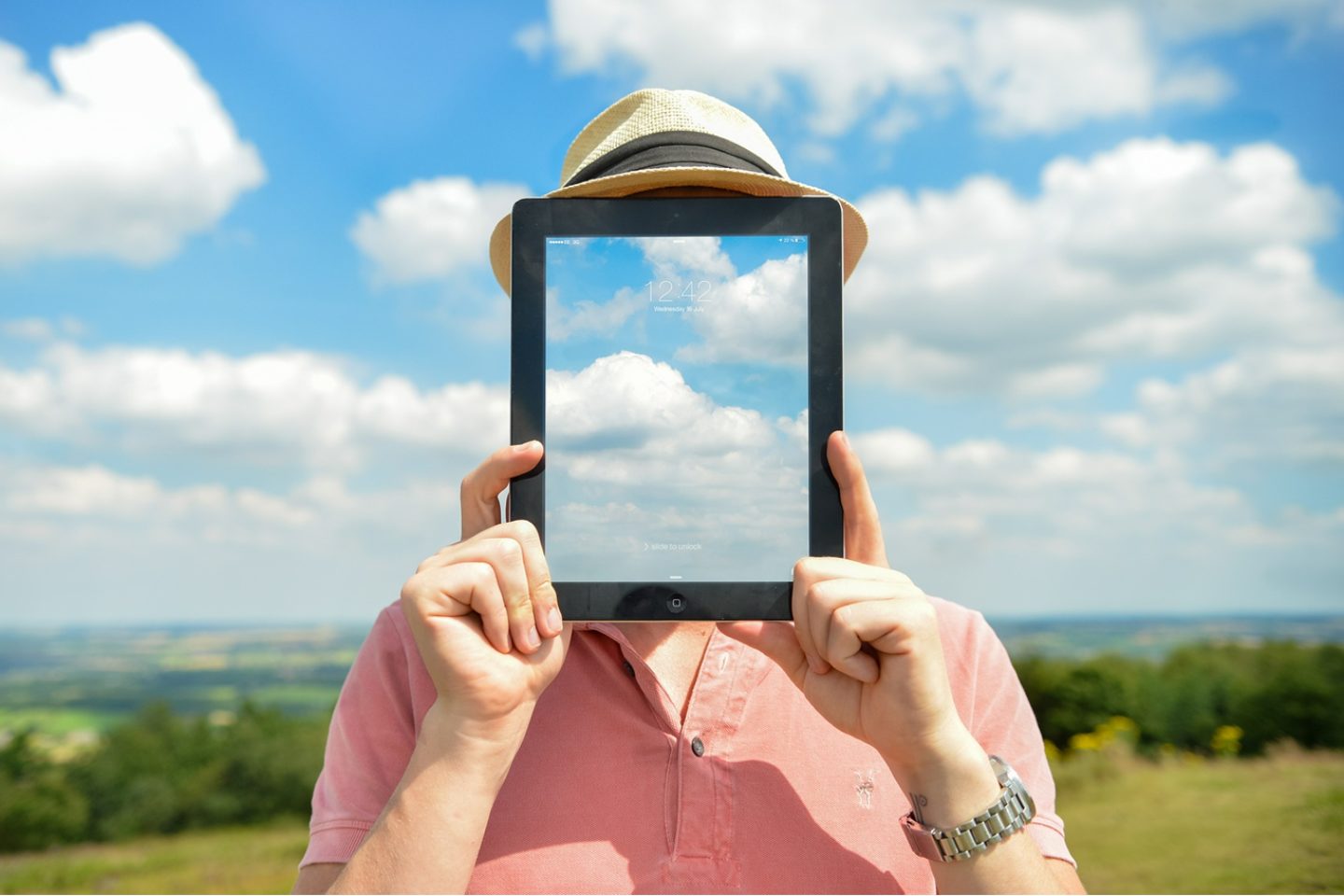 eMedia Blog with a man wearing a straw hat and pink shirt while holding up an iPad that has the same blue cloudy sky as the one behind him.