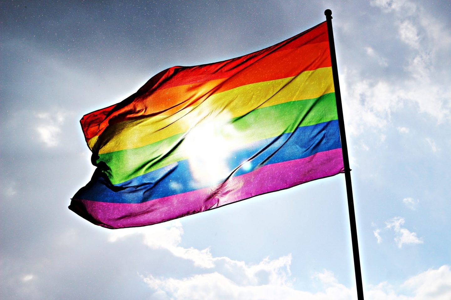 Pride flag on a flag pole in front of the sun on a blue sky with clouds.