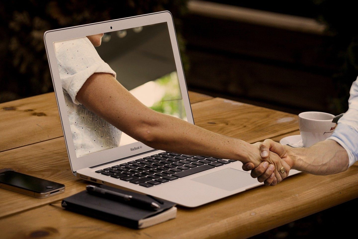 A MacBook Air is on a wooden table. Surrounding the MacBook Air is an iPhone, a notebook with a pen, and a coffee mug. The MacBook Air is open, and its screen has a person's arm coming out of it to shake the hand of the person sitting at the table.