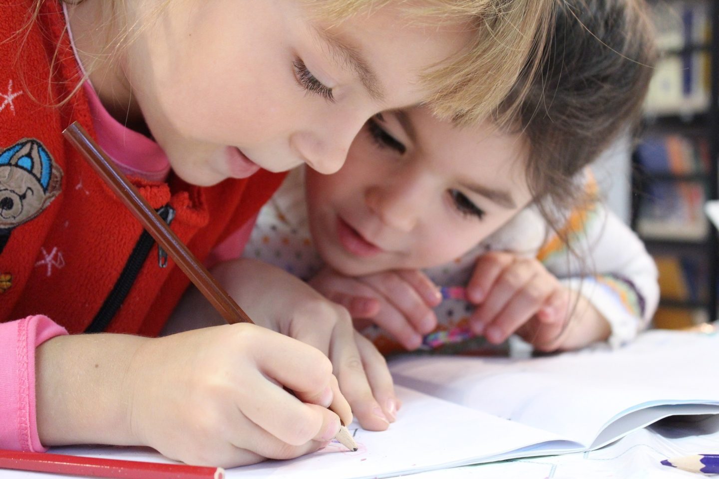 Two young girls are leaning over a book. The girl closest to the view is coloring the page with a brown colored pencil. The girl farthest from the view watches the girl color on the page.