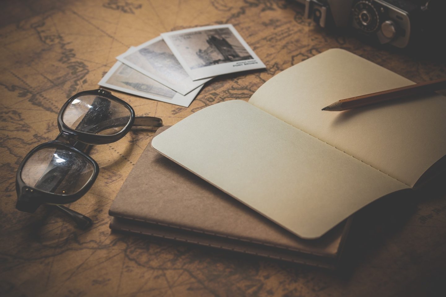 A view of a blank journal with a pencil on top of it. Below the open journal is a closed journal that sits on a desk with a map opened. There are also three black-and-white images, a camera, and a pair of black-rimmed glasses.