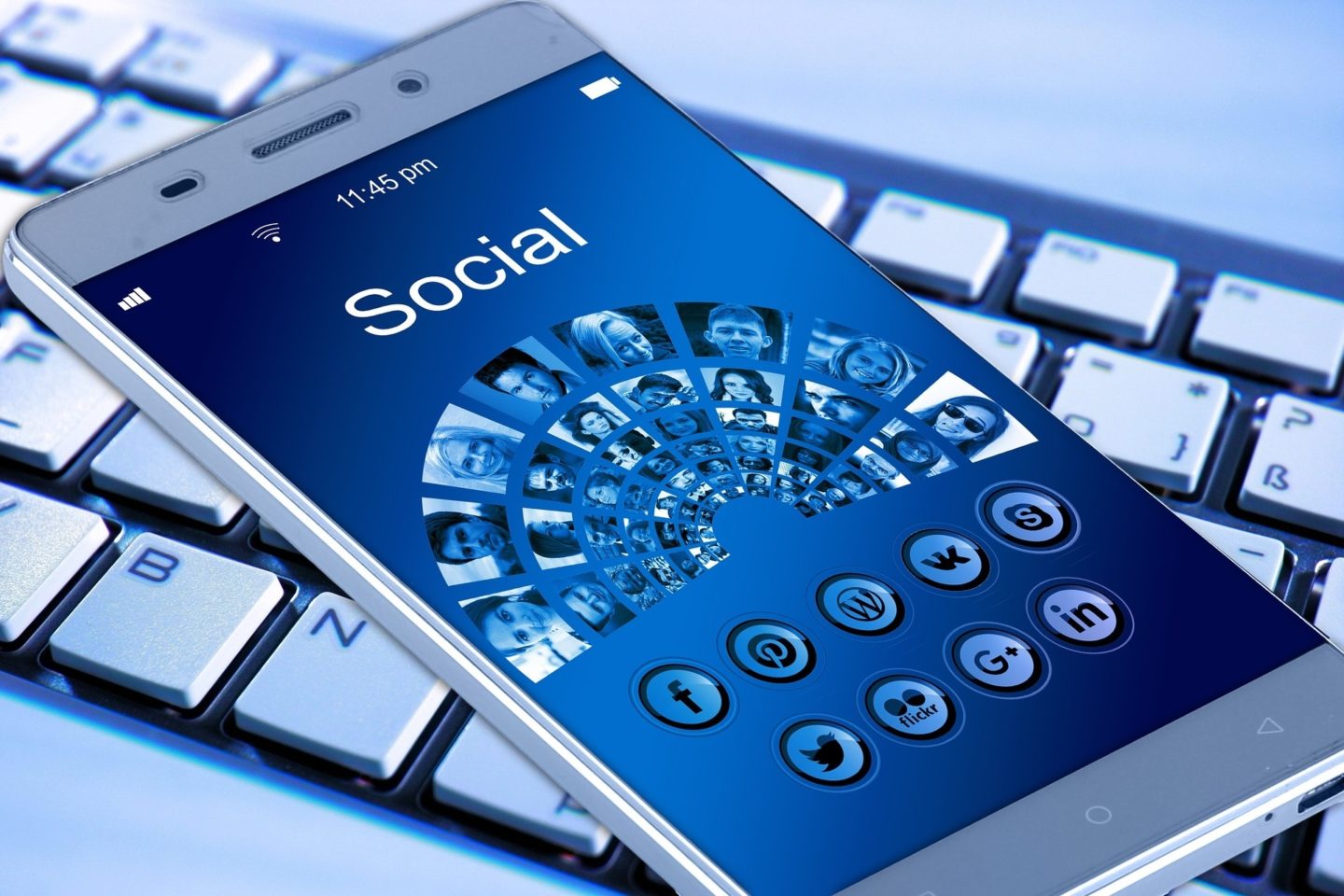 A view of a smartphone over a keyboard. On the screen is a blue wallpaper that reads "Social" and shows many profile photos and apps along the bottom of the screen. The apps includes logos of Facebook, Pinterest, WordPress, VK, Skype, Twitter, Flickr, Google Plus, and LinkedIn.