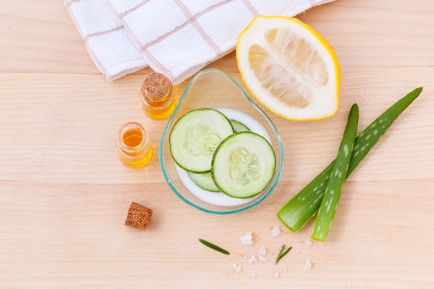 Natural skin care products are being made in a glass dish. In the dish is a cucumber and a bit of white lotion. Outside of the glass are two small glass bottles with yellow oil inside. One has its cork on and the other is open with its cork lying next to it. There is also a half-cut lemon, two strands of aloe vera, and a microfiber towel on the wooden table.