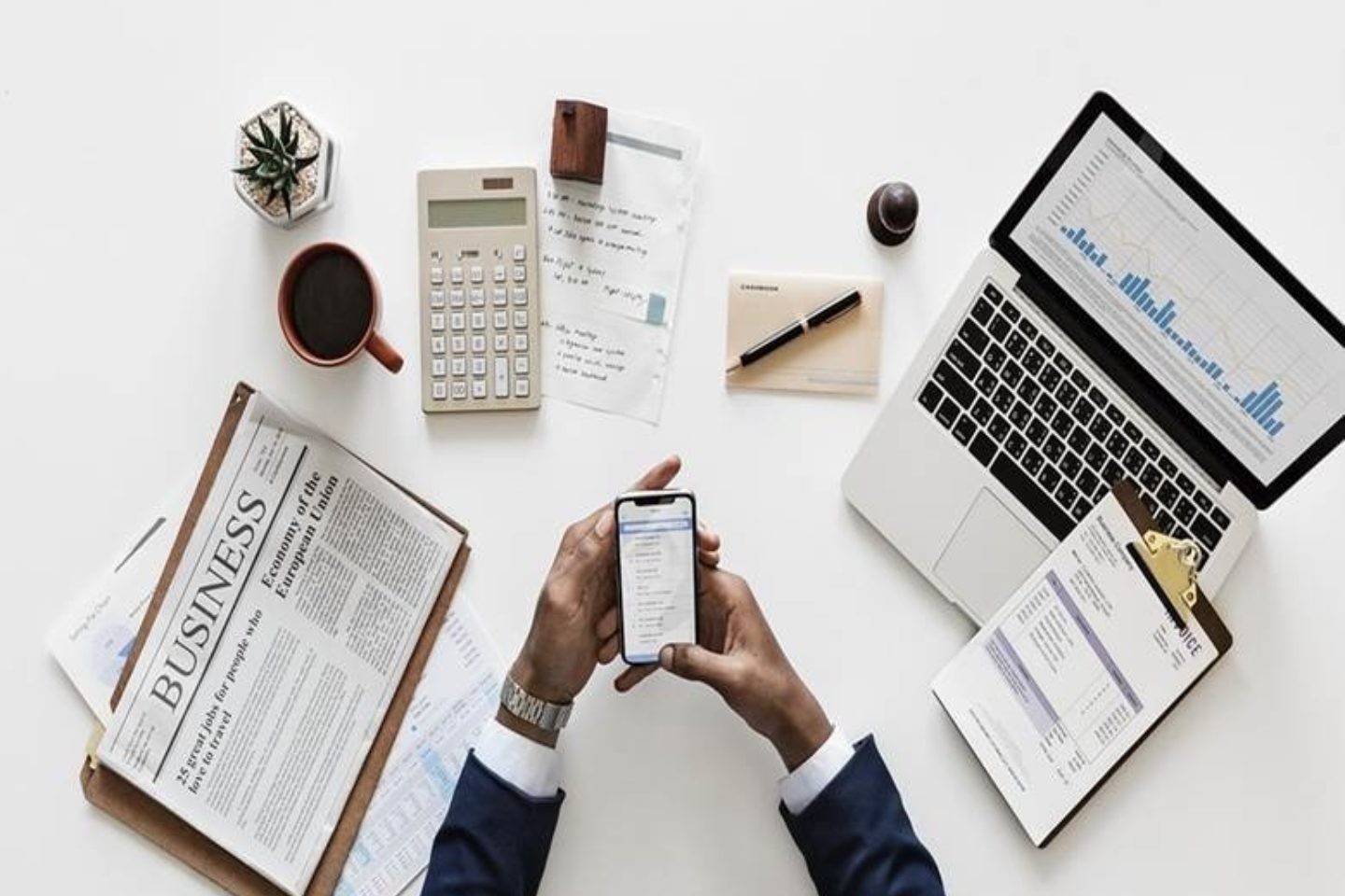 A man holds his iPhone while being surrounded by various business materials. To his left is a Business newspaper and a mug with coffee in it. Straight ahead of him on the table is a calculate, a small air plant, a note pad, a stamper, and a paper with writing on it. To the right of the man is a computer with an excel sheet open to a bar graph, as well as a clipboard with an invoice paper attached to it.