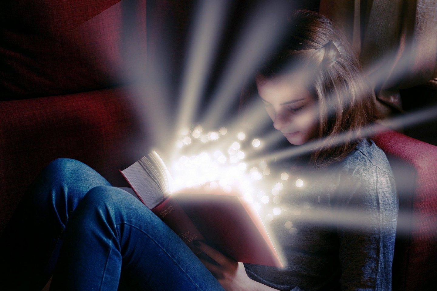 A seated girl opens a book to read that lights up the space with sparkles and light beams.