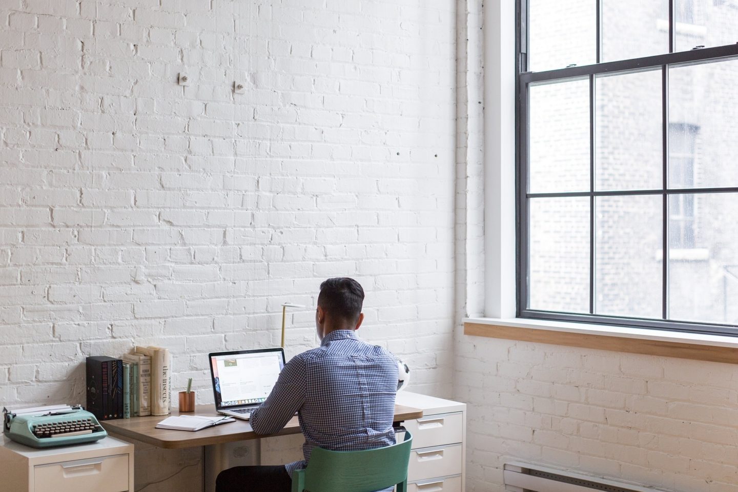 A man sits at his desk working on his laptop. To the left of him is a teal typewriter, several books lined up against the wall, and a notebook he is using while he works. He faces a white brick wall in a large room. To the right of him is a large paneled glass window looking out at another brick building.