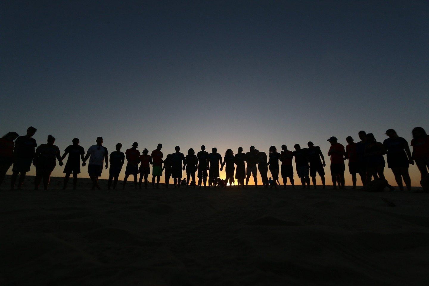 A large group of people form a semi-circle while some hold hands, and stand side by side. Behind them is a sunset view, so their bodies are all in silouette.