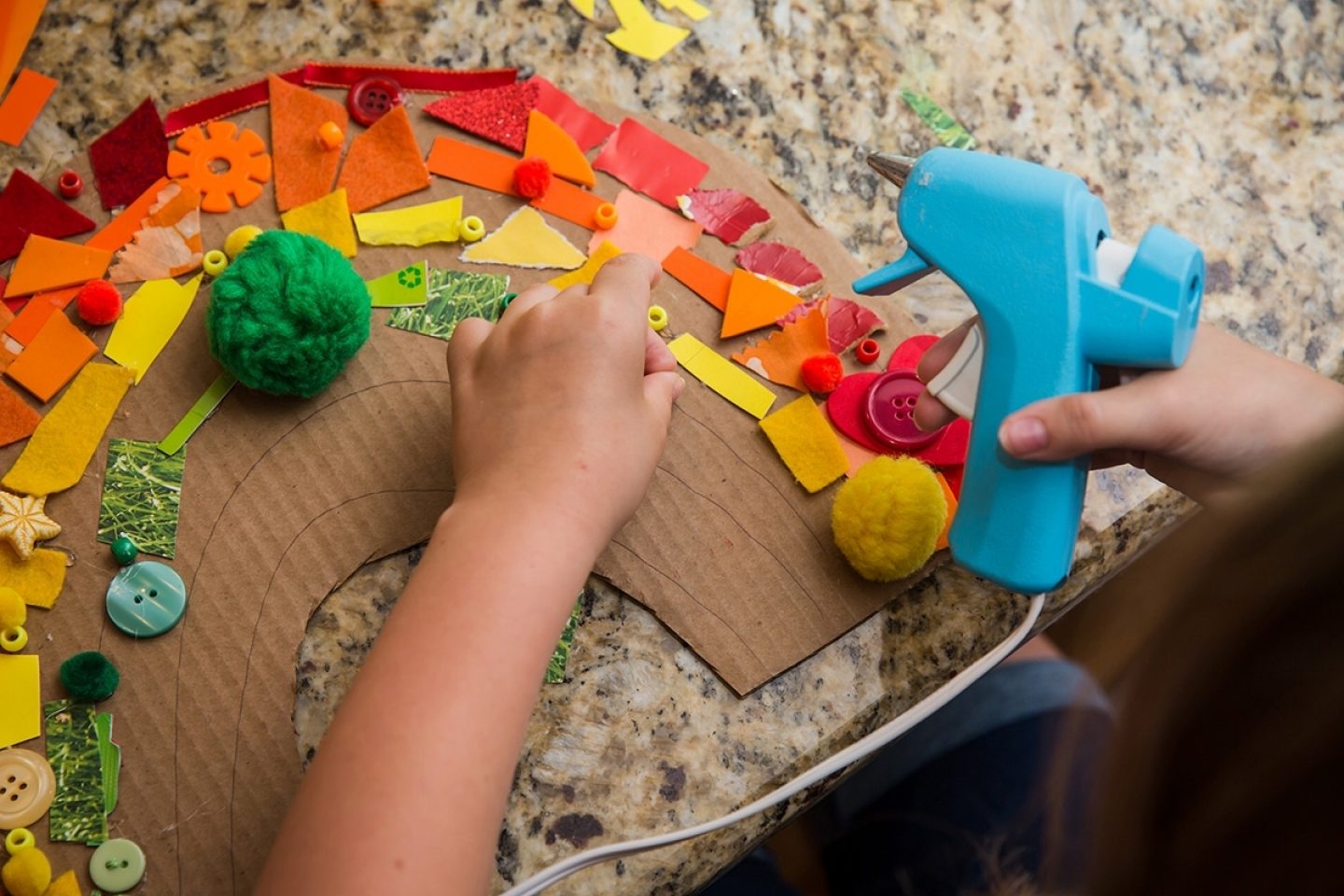 A child's hands are seen making a rainbow collage on a cardboard base with a hot glue gun.
