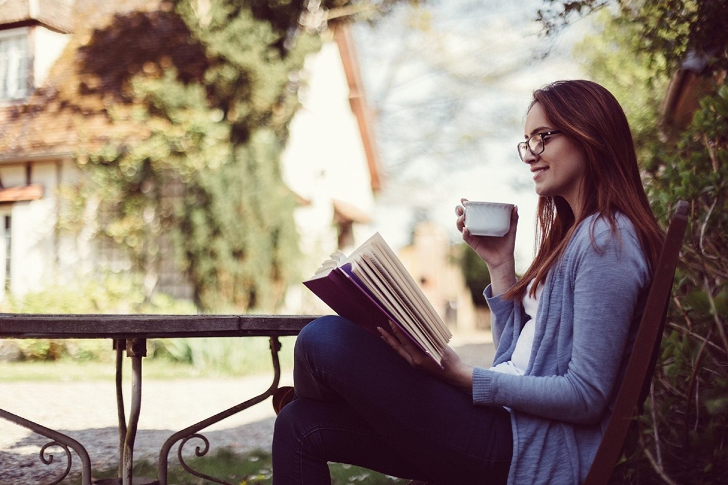 A woman drinking a cup of coffee outside is smiling while reading her book.