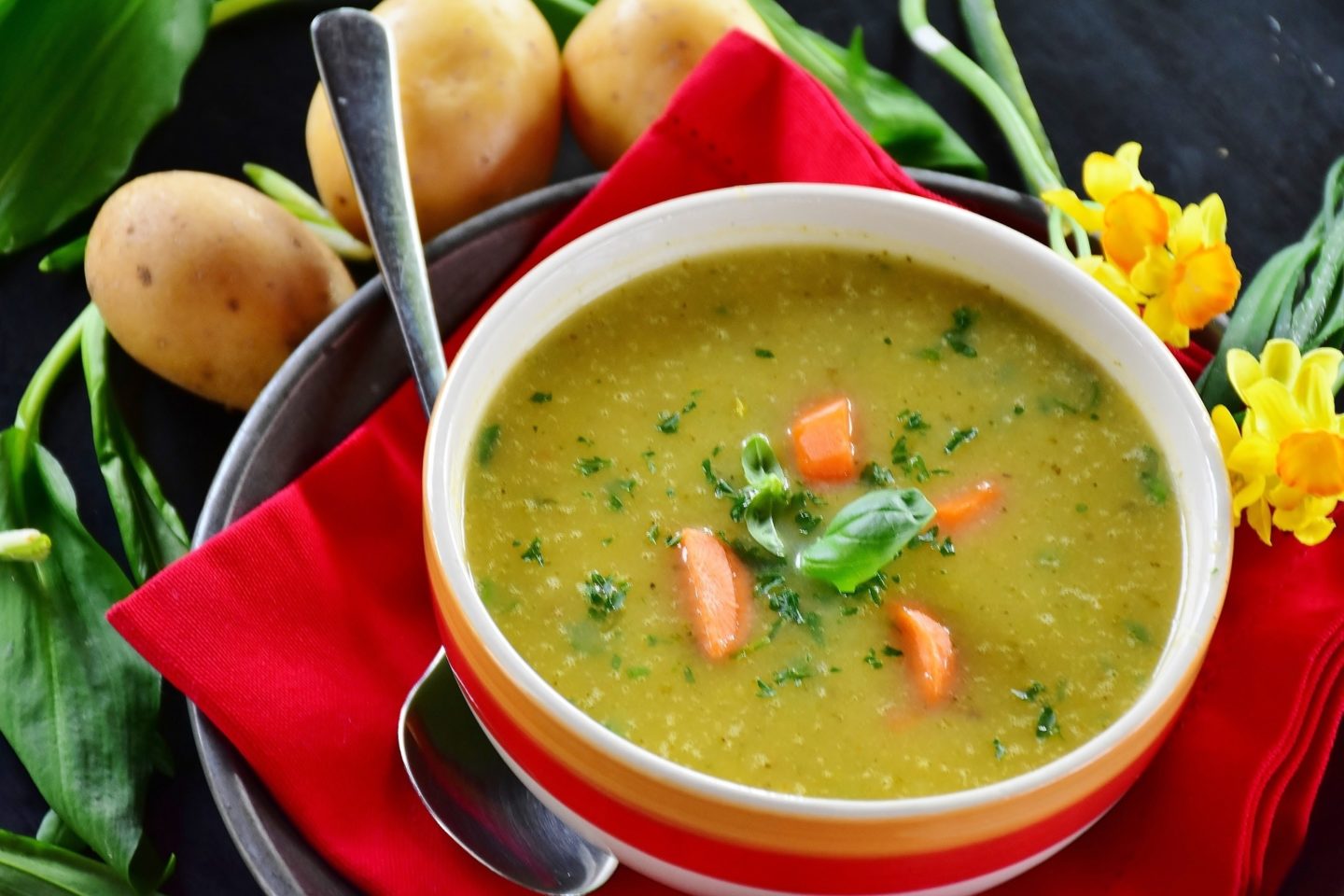 A bowl of soup sits on top of an orange napkin and a dark tray on a table surrounded by potatoes, plant greens, and yellow flowers.