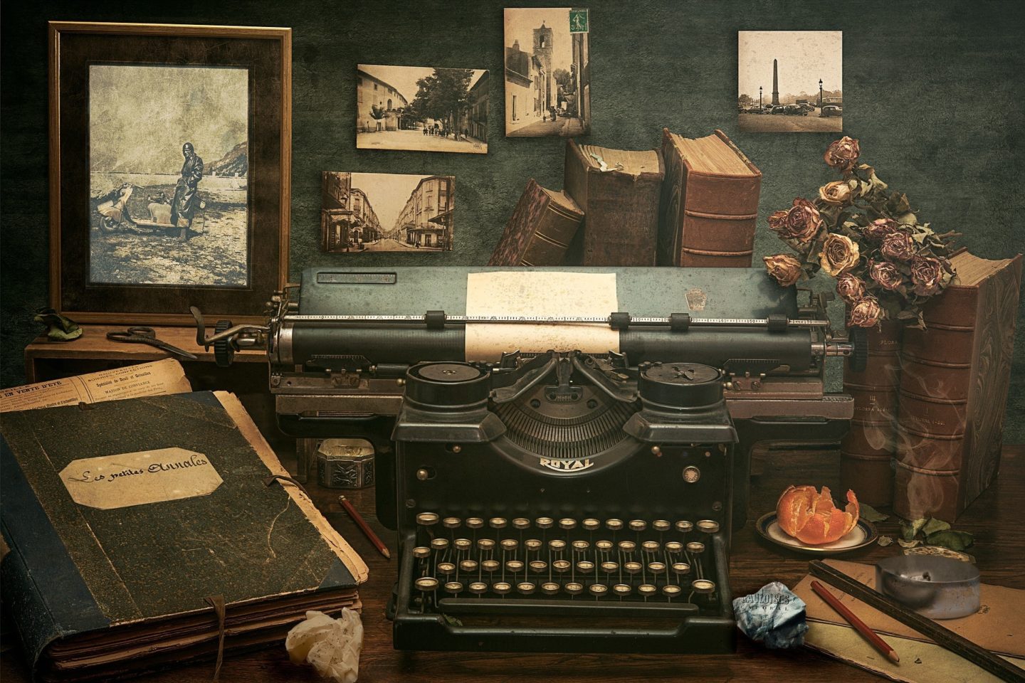 A vintage typewriter sits on a wooden desk. Surrounding the typewriter are various old items. These items include a bundle of dried roses that sit on top of two old books to the right of the typewriter. Behind the typewriter are three more old books, and four black-and-white photos that do not have frames, along with one larger framed black-and-white photo. On the table to the left of the typewriter is a booklet that appears to have newspaper clippings sticking out of the top and side. There are also two red pencils, two crumpled-up papers, an opened orange, and an ashtray that has a cigarette still having smoke coming out of it.