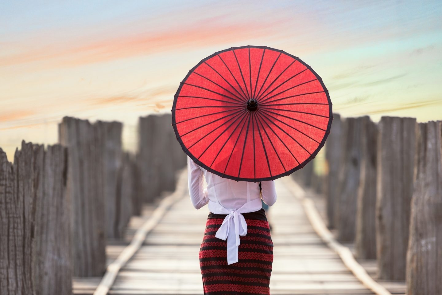 A woman walks on a wooden boardwalk with a red umbrella covering the back of her head and upper body.